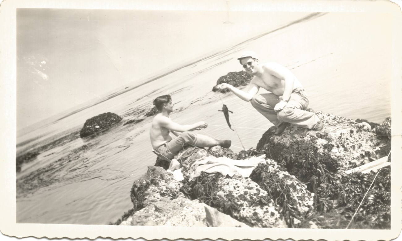 Black and white photo of two shirtless men, one holding a fish on a line, on rocks, at the edge of water.