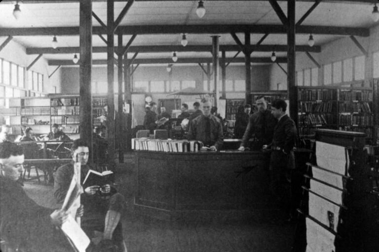 Several men sit around table reading while two stand at a large desk in the middle of the room