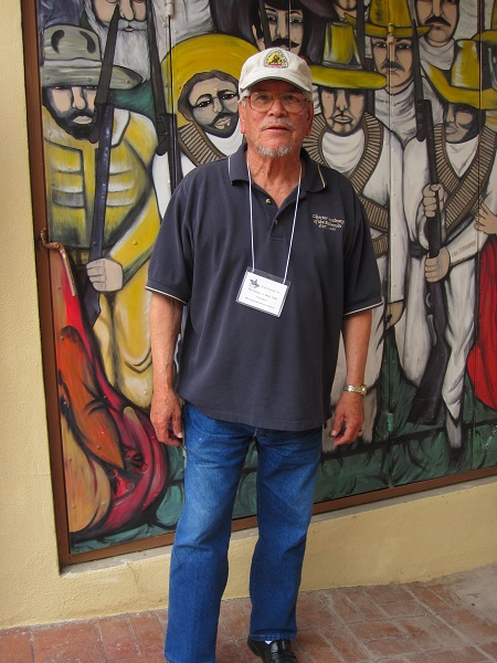 Male with a baseball cap stands in front of a painted mural on a wall. 