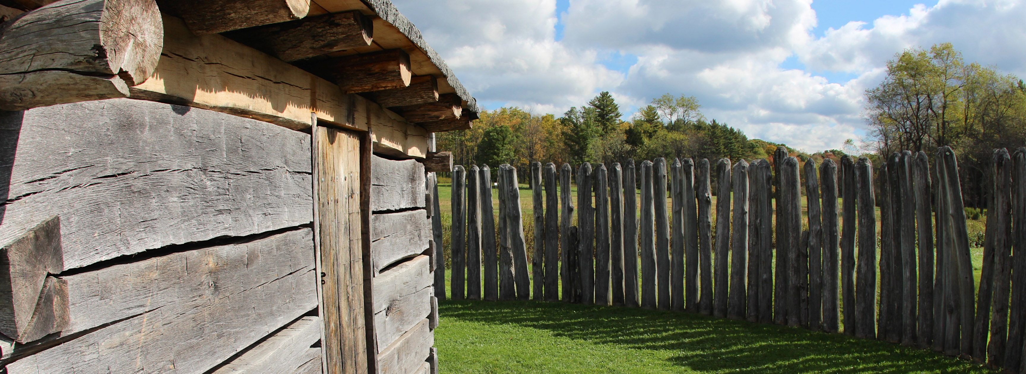 View of the side of a cabin and a post fence under a blue sky.