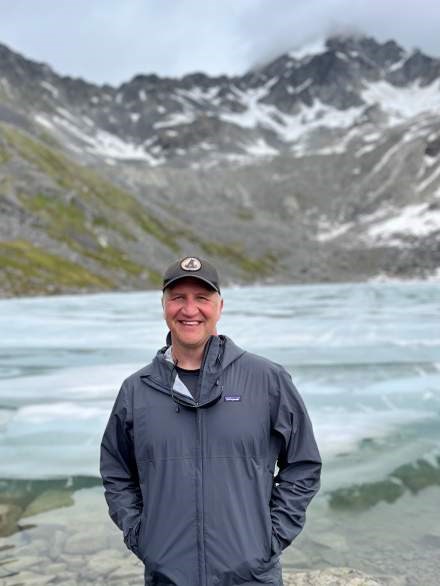 A person smiling standing in front of an alpine lake in the mountains.