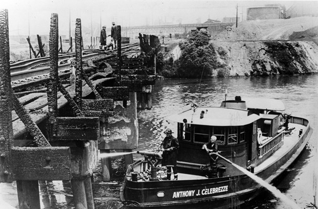 A black-and-white photo shows two firefighters spraying water from their boat up onto a burnt railroad bridge.