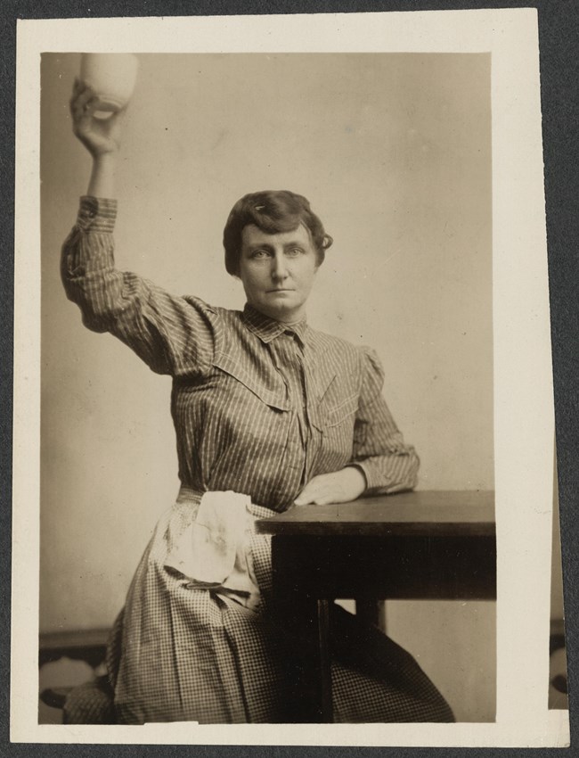 Woman wearing a prison uniform, seated with one arm raised holding a cup.