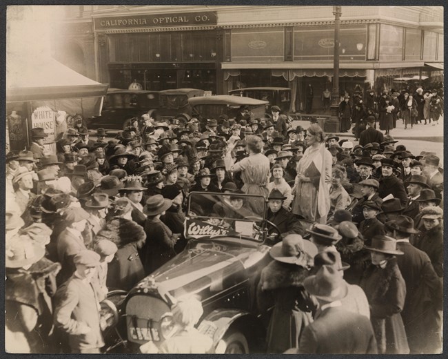 A crowd of people surround a car; two of the women in it are standing and one is speaking.