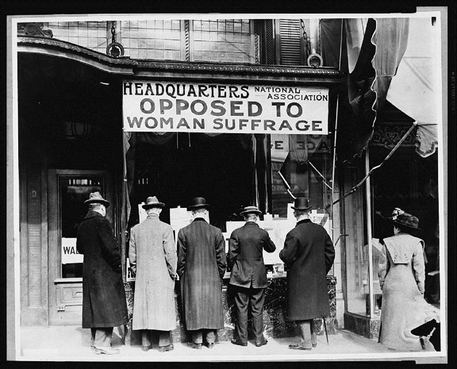 Men standing with their backs to camera under sign opposing women's suffrage. Library of Congress.