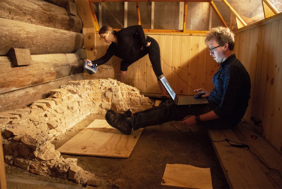 A man sits in a wooden boxy space with a laptop on his knees and feet near a crumbled brick hearth.
