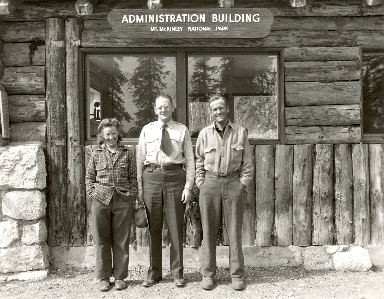 two men and a woman standing outside a log cabin with a sign that reads administration building