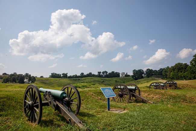 Several Civil War Cannons placed near hilltop looking out at rolling grass covered hills. A large white memorial sits atop a hill far in the distance.