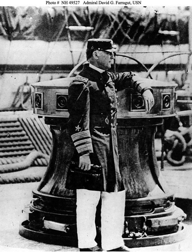 Black and white: A man is standing on a deck of a ship. His uniform shirt and hat are dark. His pants are white.