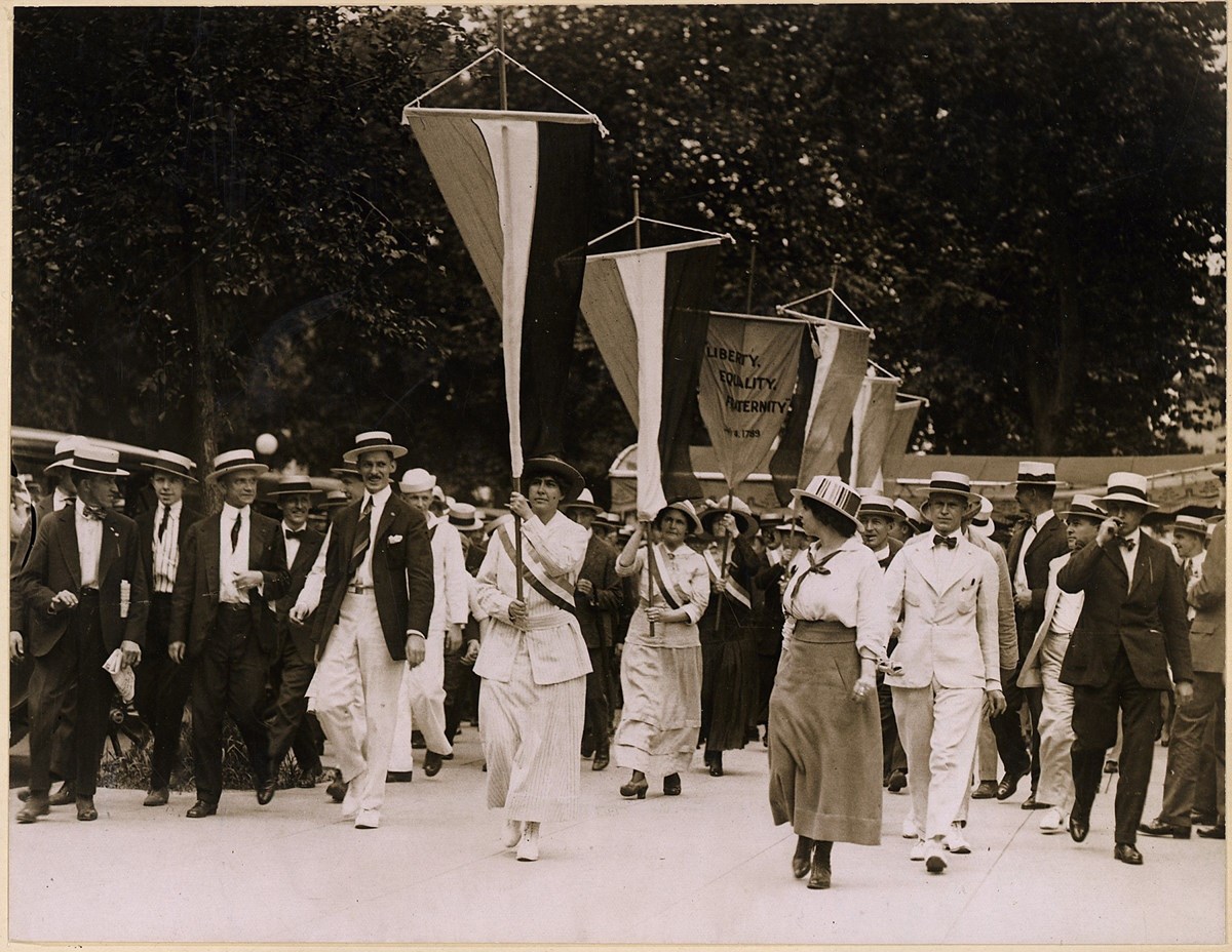 Suffragists march in a parade. Some hold suffrage flags and one holds a banner that reads, "Liberty, Equality, Fraternity."
