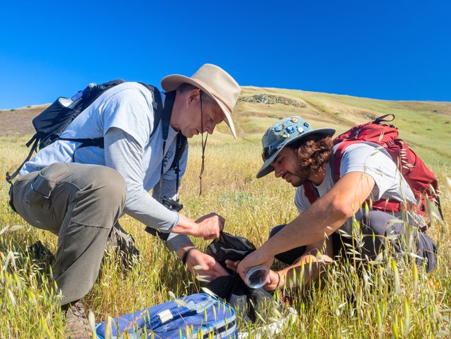 Two scientists sit on a green grassy bald on a clear blue day. The scientists are carrying backpacks and wearing brimmed hats while holding small containers presumed to hold small butterflies.
