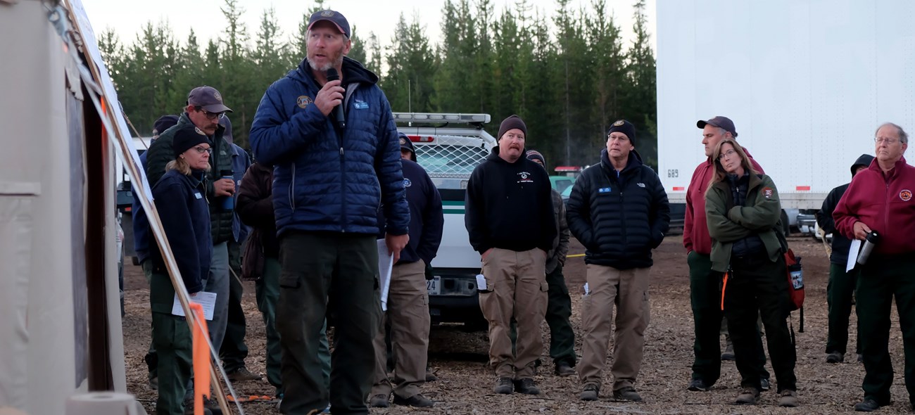 A man stands near a board in front of a group of people speaking into a microphone.