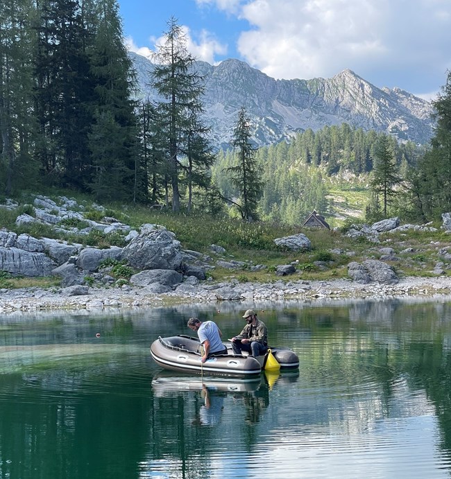 Two scientists sit in an inflatable boat studying the water in an alpine lake. In the background is a lush green forest and tall, jagged, snow-covered mountains.