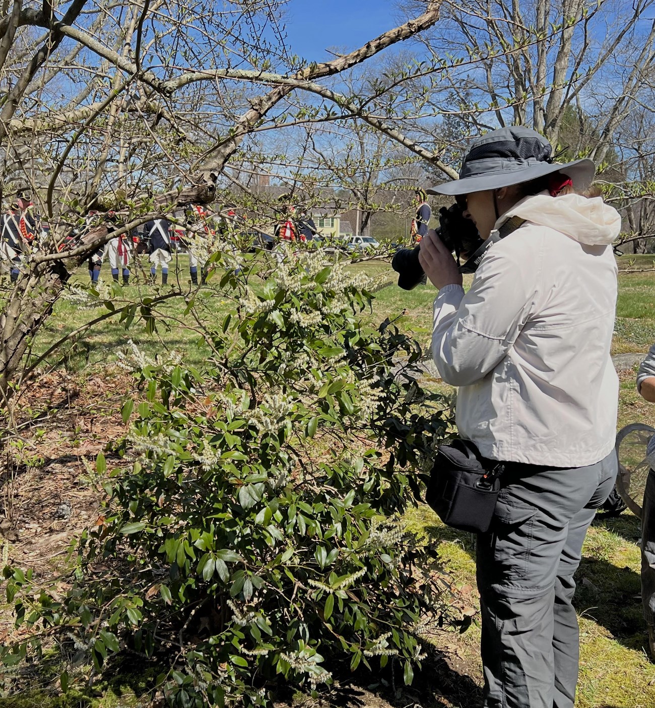 A person standing by a flowering bush holding a camera. There are Revolutionary War reenactors in the background.