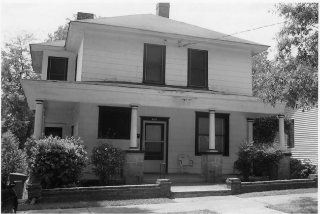 two story house with a wrapped full length porch. Steps leading up to the entrance with plants around the porch.