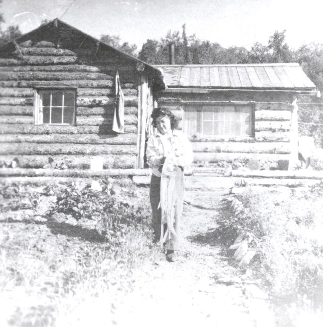 Historic black and white image of a woman standing in front of log cabin.