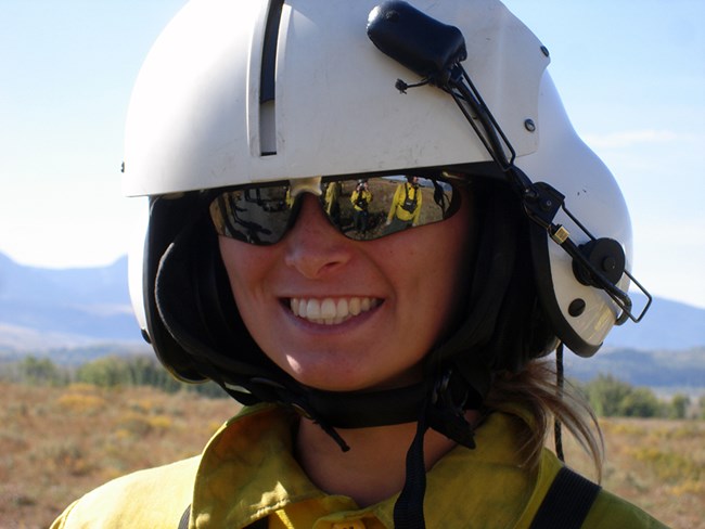 A woman in a flight helmet and sunglasses smiles.