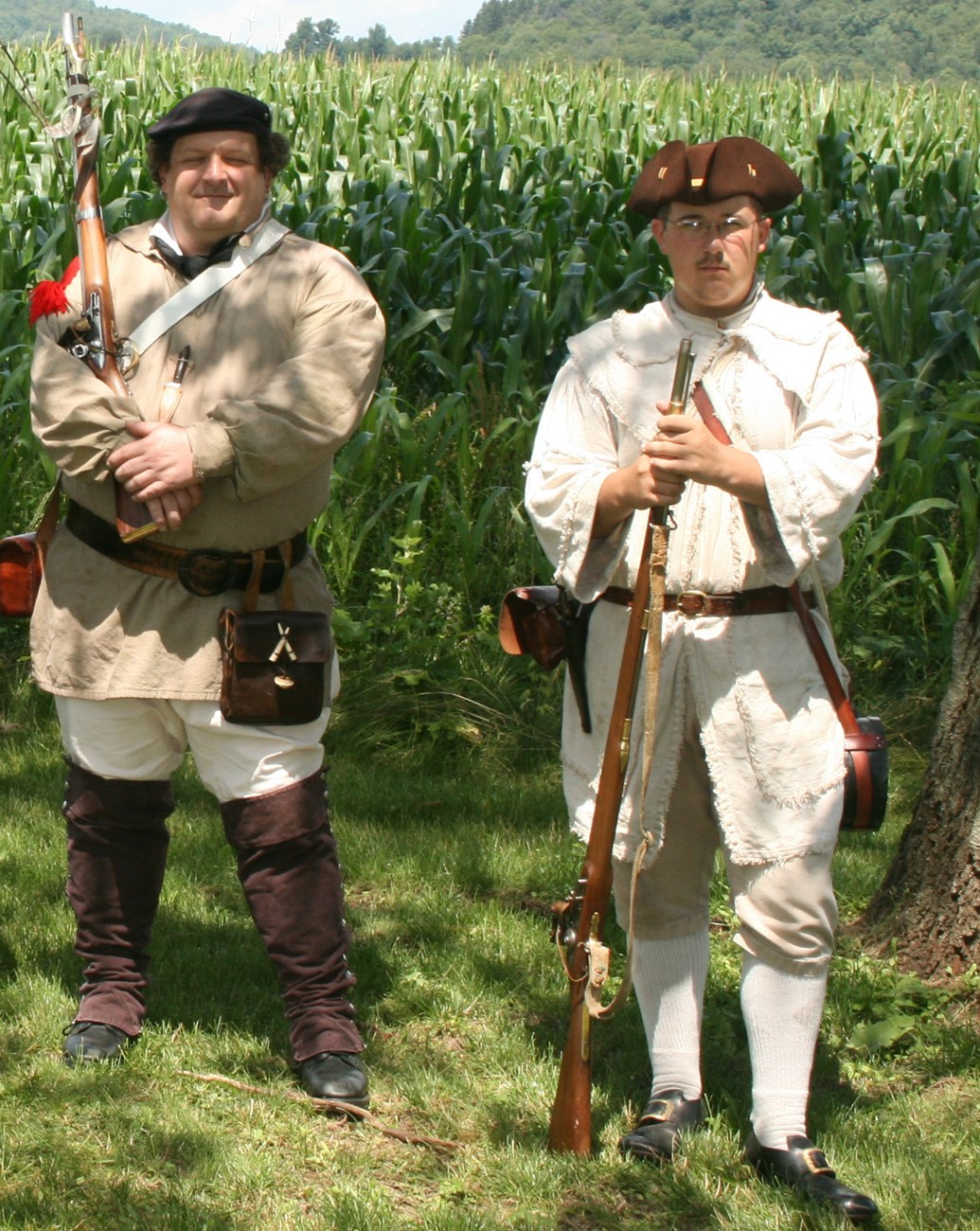 2 people in Continental Soldier uniforms stand with muskets ready under green trees.