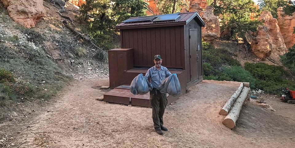 Man in park maintenance uniform stands holding two grey bags in front of brown outdoor toilet