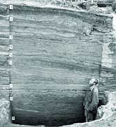Man stands in front of rock wall showing many layers of occupation at Onion Portage excavation site (1960s).