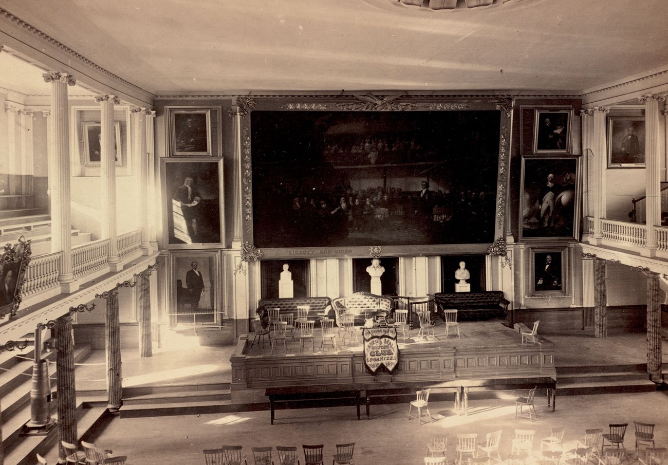 view from gallery of Faneuil Hall, an open hall with a stage and large paintings on the walls.