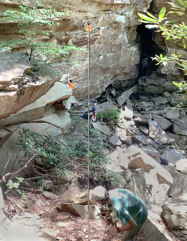 A person is next to a long pole with an orange circle at the top which is set up next to a cliff face where there are two people climbing.