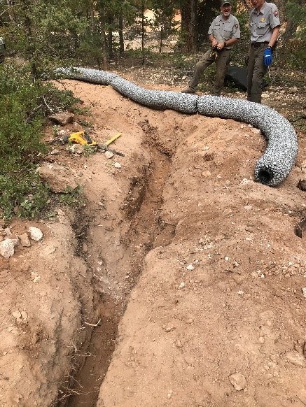 A silver tube-shaped drain sitting at the edge of a trench built for its burial. Two maintenance workers stand by to bury it.