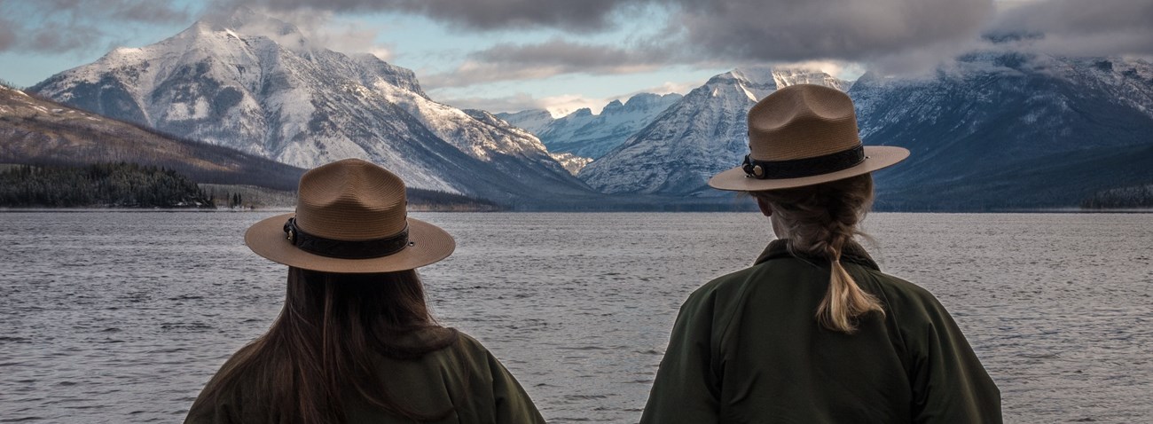Back of two rangers in flat hats looking a lake in front of a mountain
