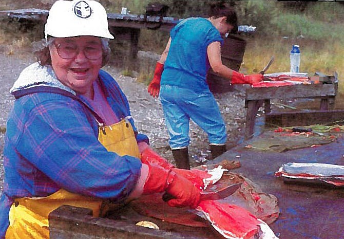 An older woman wearing a white ball-cap, blue jacket and waterproof, yellow apron cutting fish on a wet wooden table.