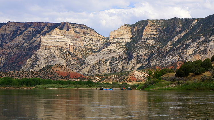 The Green River flows below beige, brown, and red cliffs with rafters in the distance.