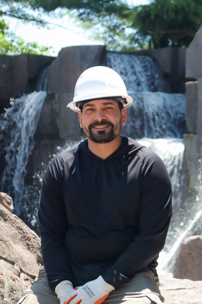 A portrait of Elvin Diaz-Ortiz smiling while seated. He is wearing a white hard hat, a black long-sleeved hooded shirt, khaki pants, and work gloves. Large granite blocks and cascading water form the background.
