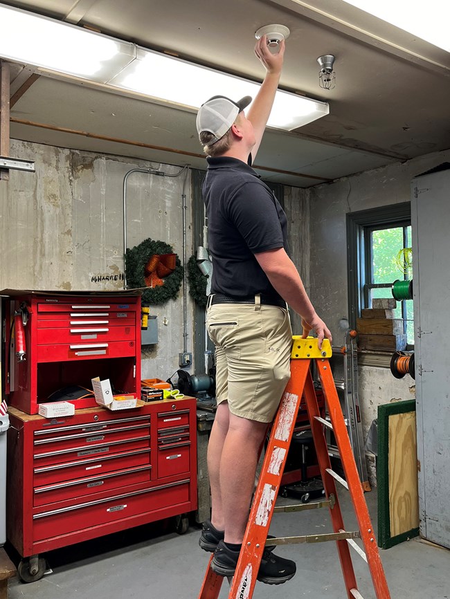 A person stands on a ladder to reach a smoke detector on the ceiling.