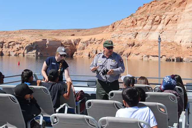 A park ranger holding binoculars speaks to a group of children on the top of a exposed deck boat. The boat navigates a narrow channel with tall red canyon walls.
