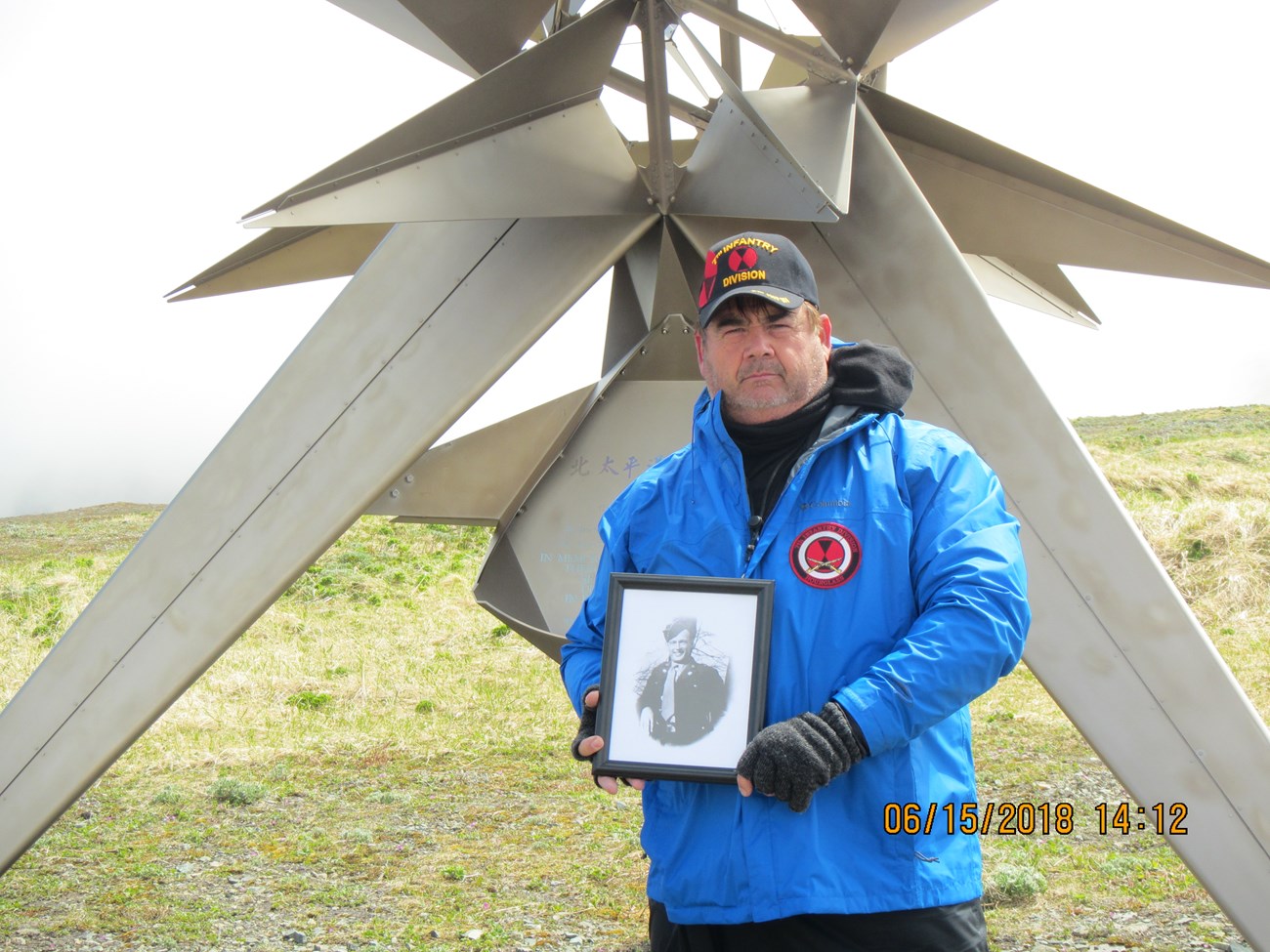 Man in blue raincoat holding black and white portrait of man in uniform, in front of large metal sculpture on top of a hill.