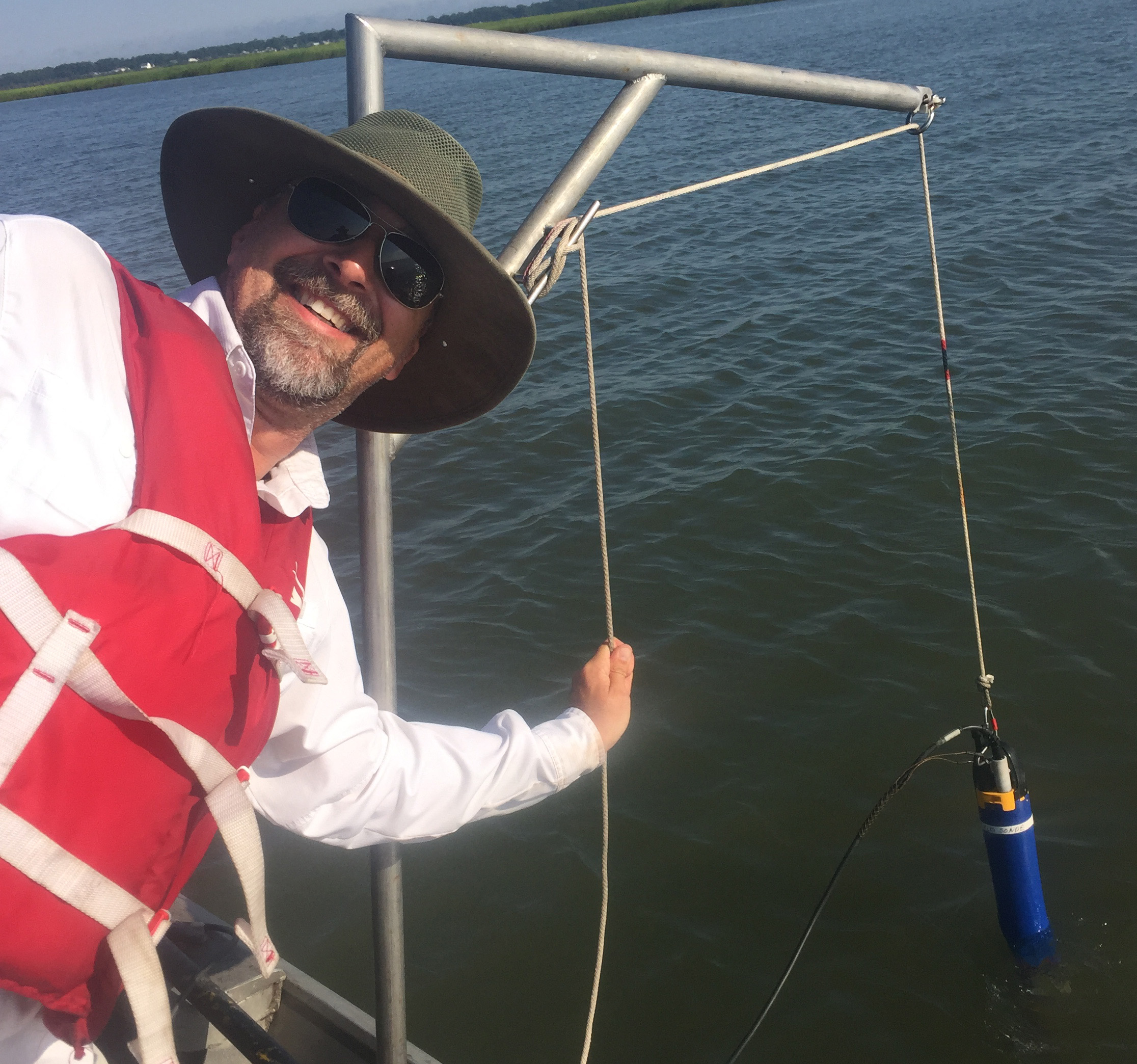 Man smiles at camera while a datasonde is lowered into the water behind him.