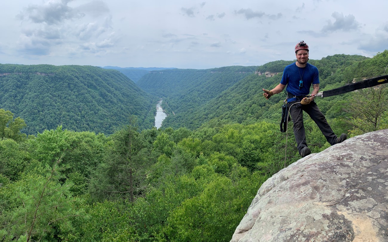 A researcher giving a thumbs up who is harnessed and connected to a rope at the edge of a cliff overlooking a lush valley.
