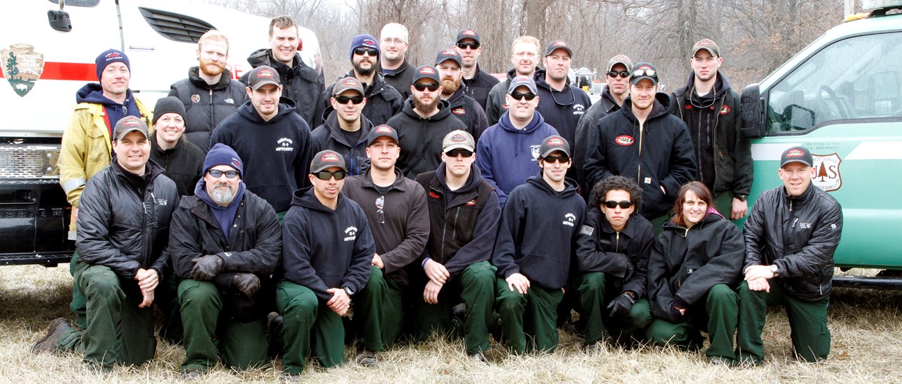 A group of people stand and kneel between a vehicle from the National Park Service and a vehicle from the U.S. Forest Service.