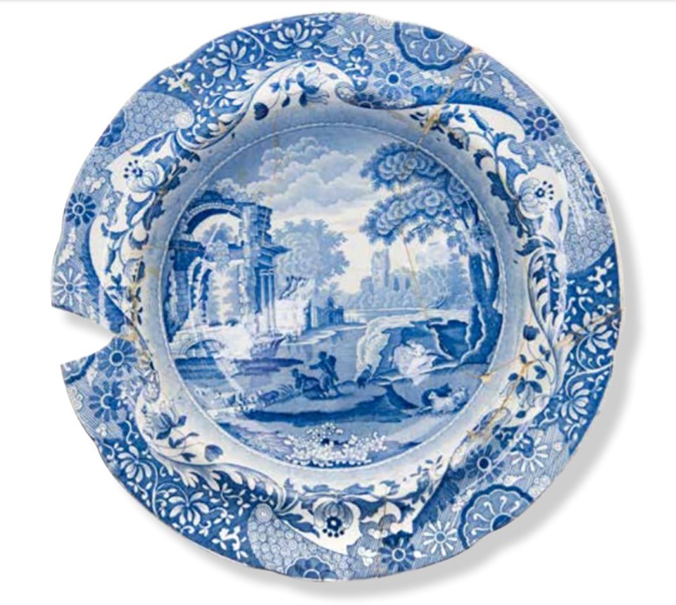 A plate with a blue transferprinted design showing buildings, flowers, people, and animals. The plate is broken and repaired, with some small fragments missing.