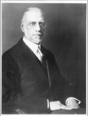 An older man seated for a formal portrait in a suit and tie. The portrait is in black and white.