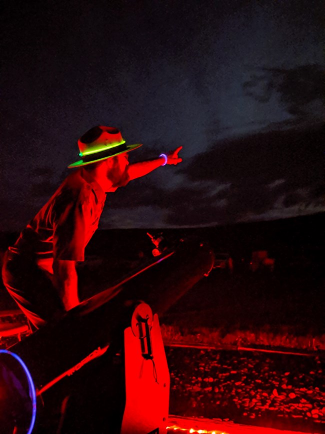 Glow from red lights illuminates a telescope and park ranger, both directed towards the night sky.