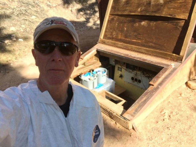 Man in white cleaning suit takes a selfie in front of open wooden compartment attached to outdoor toilet