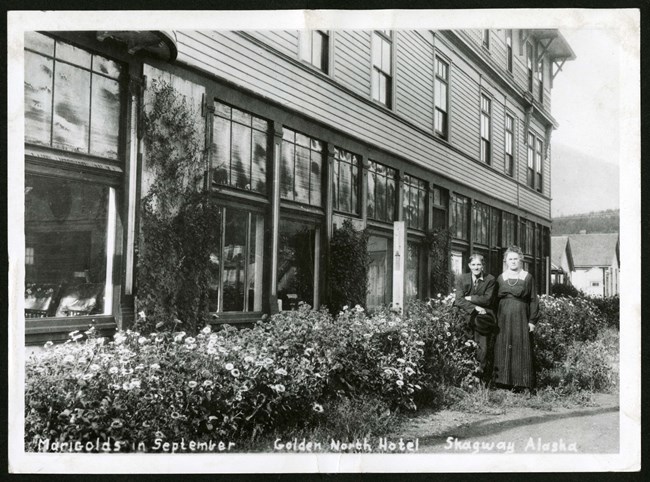 Older man and woman among marigold flowers in front of a hotel.