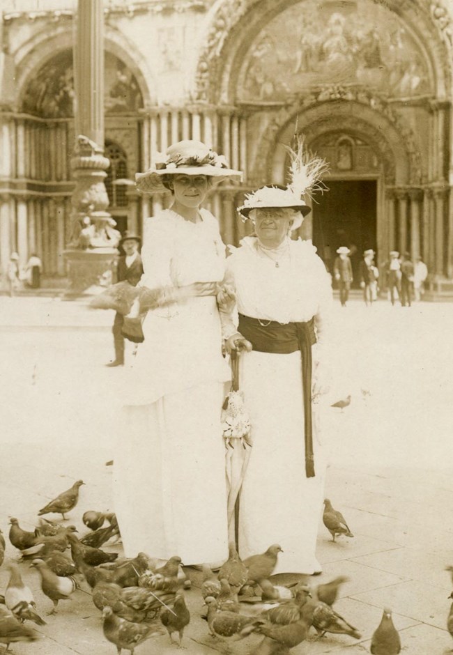 Two women in white dresses and hats standing in public square surrounded by pigeons.