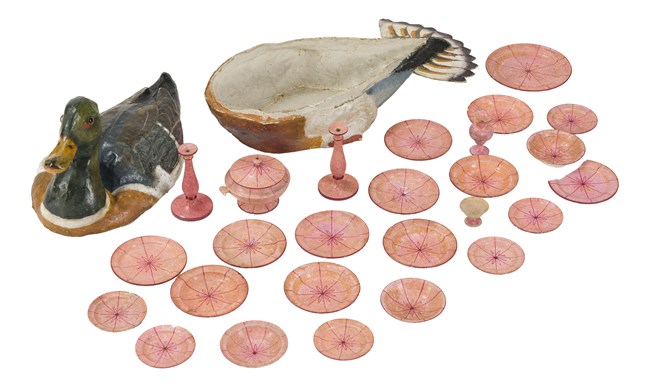 A paper mâché box in the shape of a duck holds a pink dinner doll set, featuring small wooden plates, bowls, tureen, and candlesticks