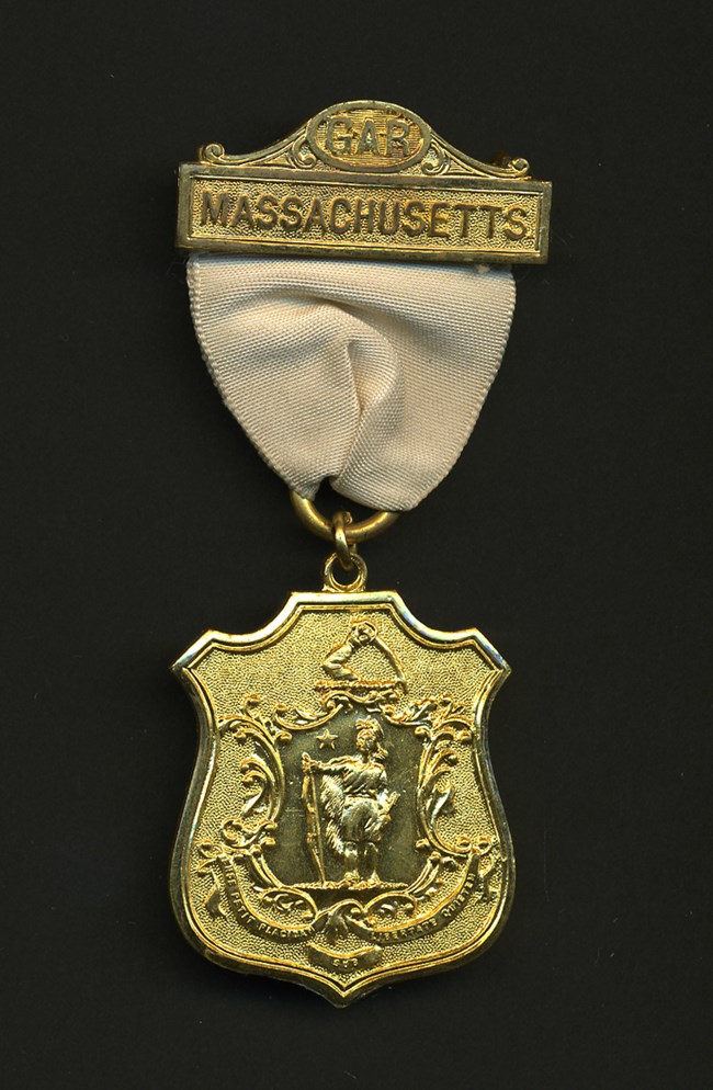 Gold-colored metal badge on off-white ribbon with seal of Massachusetts and text GAR Massachusetts on top bar