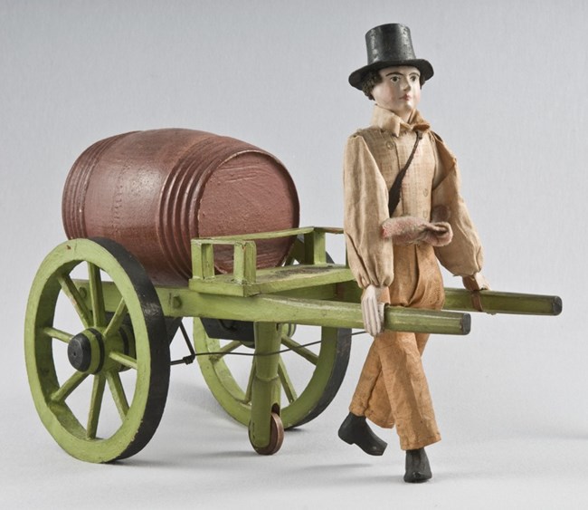 A toy featuring a man with a top hat pulling a wheelbarrow holding a barrel of buttermilk