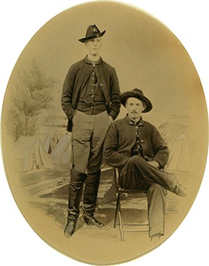 Studio portrait of two young men in Civil War uniform in front of painted backdrop with camp scene