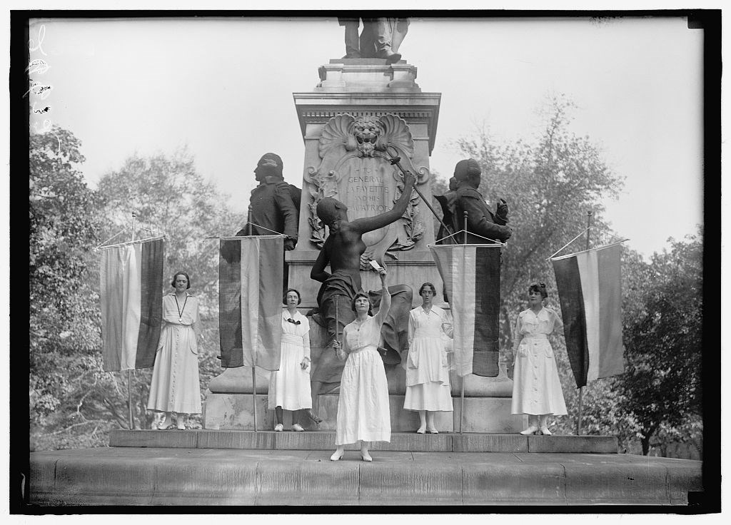 Women holding suffrage flags in front of a statue of Lafayette. The woman in the center holds a torch and a piece of paper.