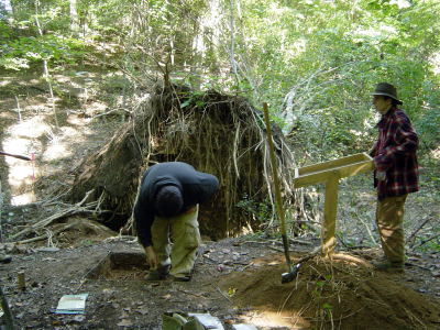 Photograph of two archaeologists at a dig site. The man appears to be digging while the woman appears to be holding up a wooden sign.