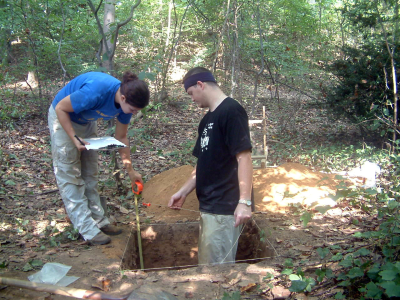 One archeologist (woman) is measuring the depth of the ditch with a measuring tape as the other (man) stands inside.